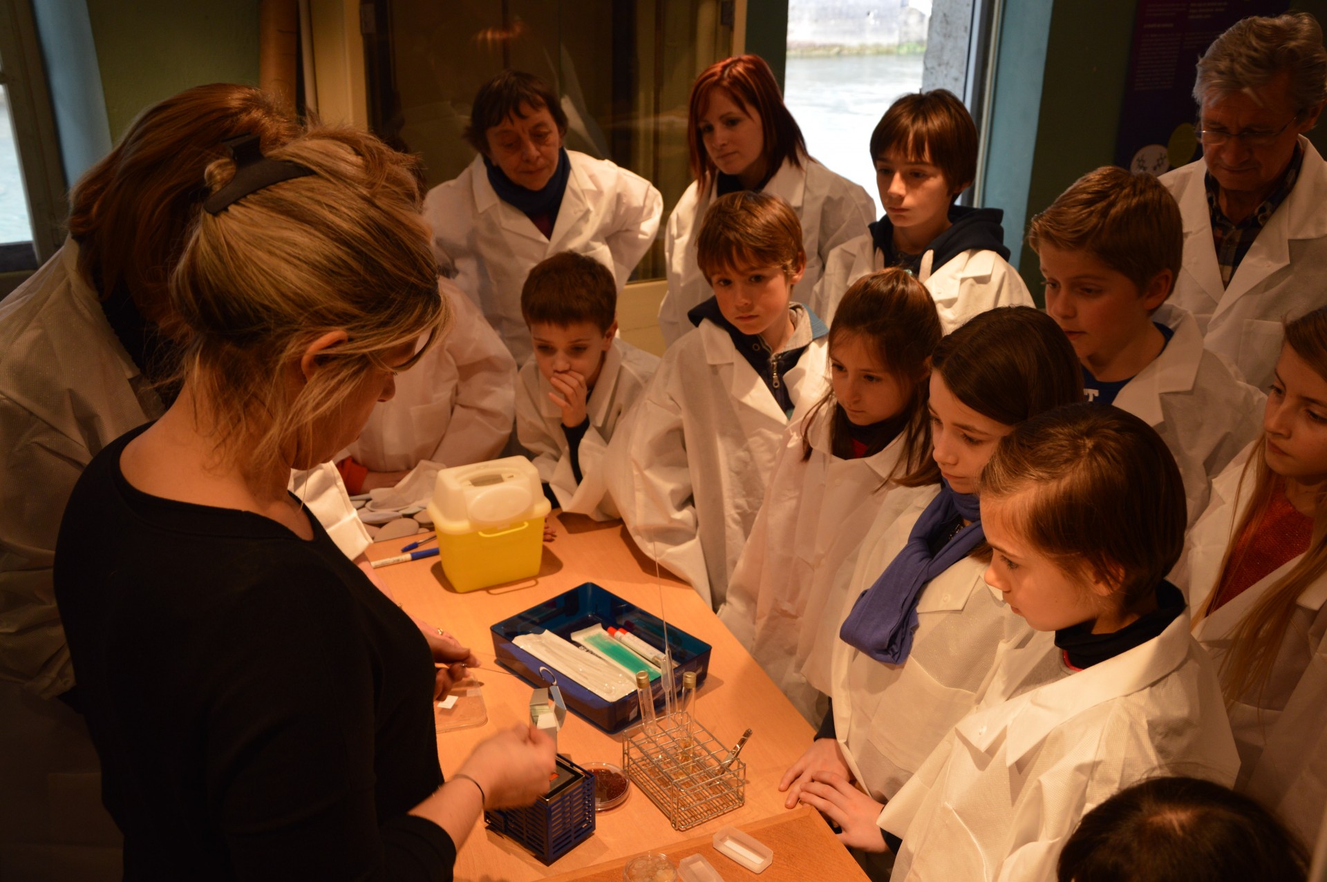 Des enfants et des adultes en blouses blanches de scientifiques sont runis autour d'une table et sont en train d'couter les explications d'une dame.