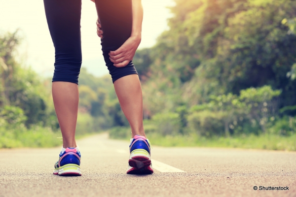 Zoom sur les jambes d'une coureuse avec une route en forêt en fond. La coureuse tient son genou, témoignant d'une douleur