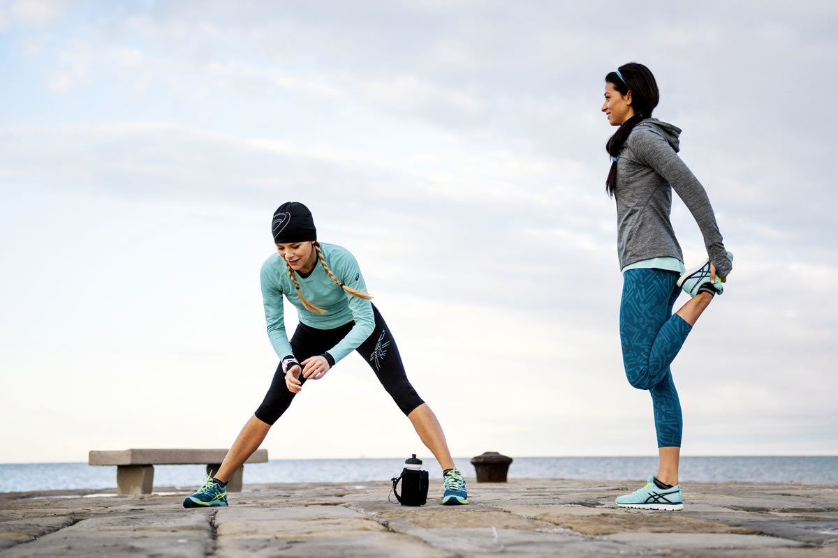 Deux femmes s'étirent après une course, l'une replie son genou contre sa cuisse, l'autre étire ses jambes.